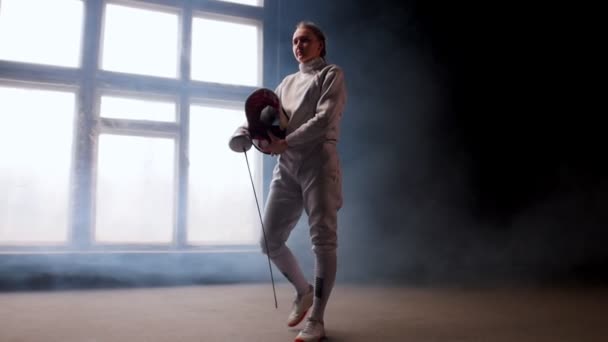 Een jonge vrouw fencer zetten op een beschermende helm en krijgt in positie - rokerige studio — Stockvideo