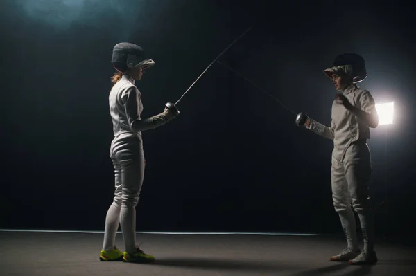 Two young women having training in a fencing duel in the smoky studio - greeting each other and starting the duel