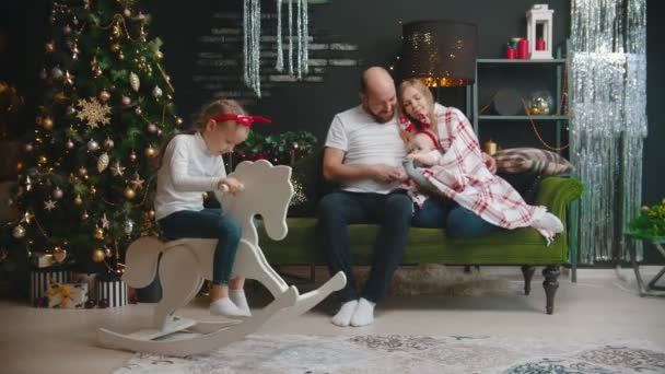 Conceito de Natal - família refrigeração no sofá na sala de estar - filha brincando no cavalo de brinquedo — Vídeo de Stock