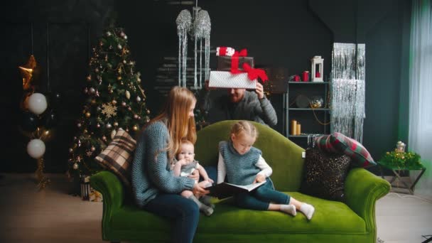 Concepto de Navidad - una madre con sus hijos sentados en el sofá - papá asomándose desde detrás del sofá y trae los regalos — Vídeos de Stock