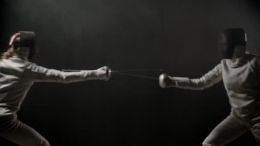 Fencing training - two young women having a duel between each other in the dark smoky studio