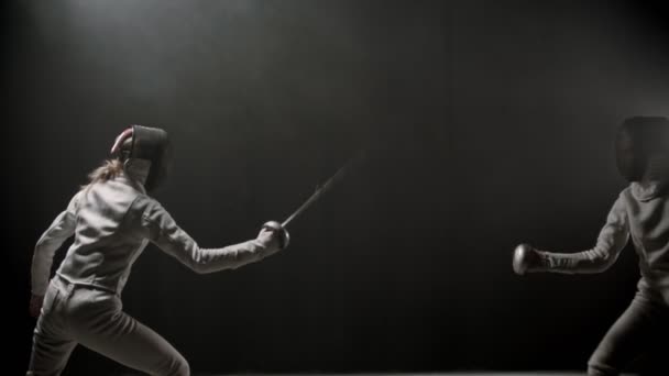 Тренування фехтування в приміщенні - дві молоді жінки мають дуель між собою в темній димній студії — стокове відео