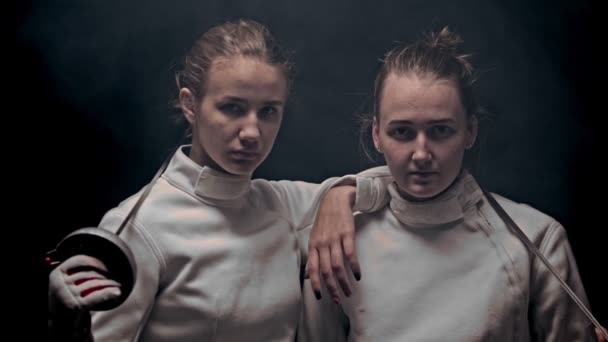 Two young women fencers standing in the dark studio - posing - raising their heads up and looking in the camera — Stock Video