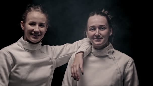 Két fiatal, mosolygó női vívó áll a stúdióban, és a kamerába néznek. — Stock videók