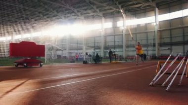 Kapalı alanda atlayan sarı gömlekli bir adam stadyumdaki barın üzerinden atlıyor.