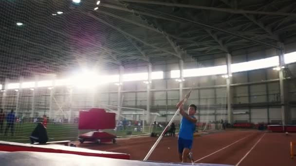 Entrenamiento de bóveda de poste en el estadio: un joven con camisa azul saltando sobre la barra — Vídeos de Stock