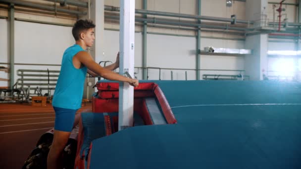 Saut à la perche - jeune homme en t-shirt bleu tourne la grue et élève la barre — Video