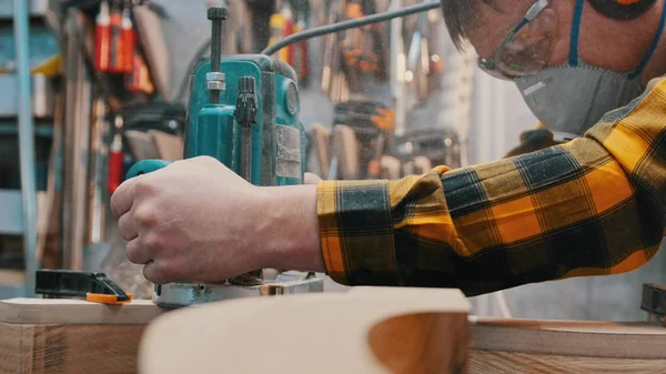 Carpintería en el interior - un carpintero pule el detalle de madera desde la parte superior del taller - un hombre con auriculares protectores y máscara —  Fotos de Stock