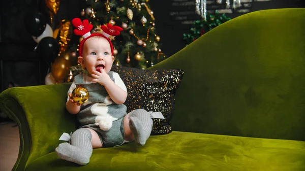 Concepto de Navidad - sonriente bebé feliz está sentado en el sofá verde y jugando con la bola de Navidad — Foto de Stock