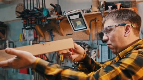 Snickeriindustrin - arbetar i verkstaden - en man träarbetare i skyddsglasögon inspektera trä detalj för brister — Stockvideo