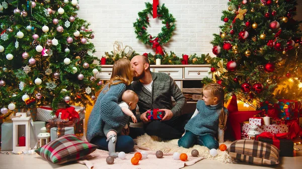 クリスマスのコンセプト-幸せな家族はリビングルームでクリスマスの設定に座ってギフトを交換します-お母さんとお父さんのキス — ストック写真