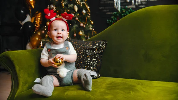 Concepto de Navidad - bebé sonriente está sentado en el sofá verde y jugando con la bola de Navidad — Foto de Stock