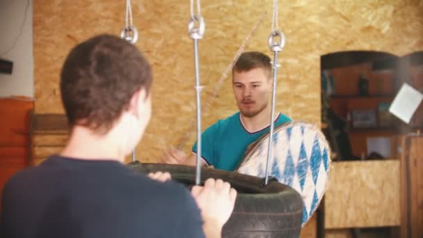 Мужчина рыцарь тренируется на мечах на связке шин в тренажерном зале - другой человек держит связку — стоковое видео