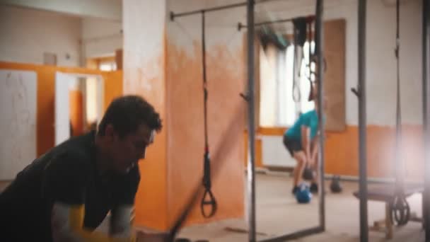 Un hombre con una camiseta negra golpeando un neumático grande con un martillo de metal en el gimnasio - otro hombre en cuclillas con peso en las manos — Vídeos de Stock