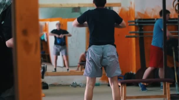 Spor eğitimi - spor salonunda egzersiz yapan erkekler - ön planda mavi şortlu bir adam elinde bir ağırlıkla çömeliyor. — Stok video