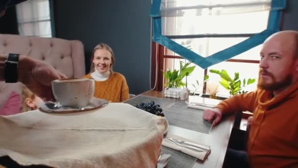 En framgångsrik familj sitter i hotellets restaurang - en servitör tar kaffekoppar till bordet — Stockvideo