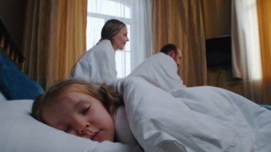 Otel odasında bir aile - yatakta uyuyan küçük bir kız - anne ve babası perdeleri açıyor ve kız uyanıyor