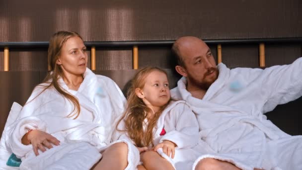 En lykkelig familie i hvite badekåper som sitter i senga og snakker med hverandre – stockvideo