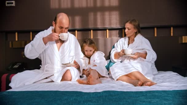 Uma família feliz bem sucedida em roupões de banho brancos sentados na cama do hotel e bebendo chá — Vídeo de Stock