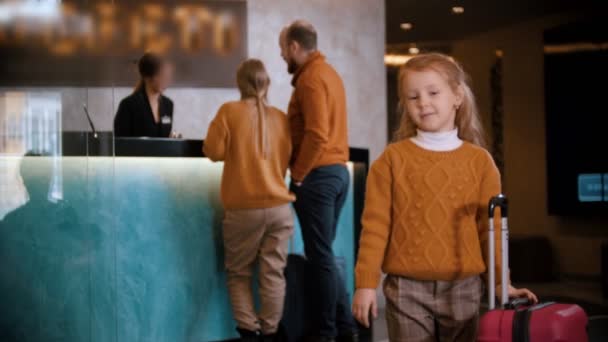 Zameldowanie rodzinne w hotelu - mała dziewczynka stojąca z walizką i czekająca na rodziców — Wideo stockowe