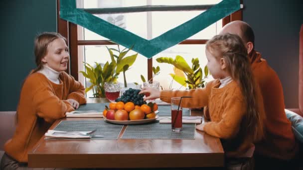 Uma família feliz sentada no restaurante do hotel - uma menina comendo uvas e alimentando seus pais — Vídeo de Stock