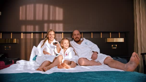 Una familia con batas blancas sentada en la cama y viendo la televisión — Vídeos de Stock