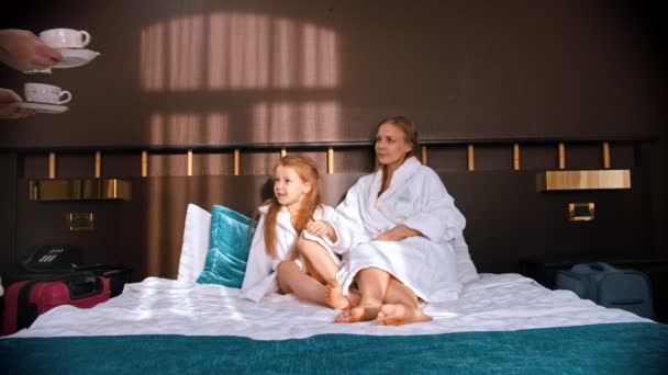 Rodzina w białych szlafrokach w pokoju hotelowym - tata przynosi filiżanki herbaty do łóżka żonie i córce — Wideo stockowe