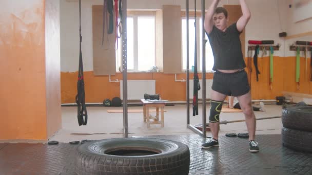 Trening sportowy w pomieszczeniach - człowiek z ochronną poduszką kolanową uderzający w dużą oponę metalowym młotkiem w siłowni — Wideo stockowe