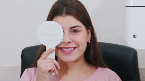 Офтальмологічне лікування - молода жінка перевіряє гостроту зору - закриває очі щитом для очей і каже, що говорить проекція — стокове відео