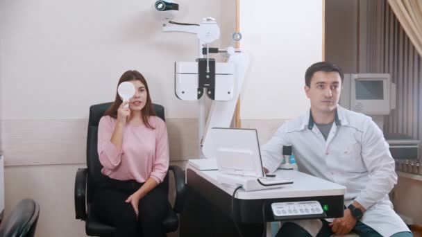 Tratamiento oftalmológico - mujer joven comprobando su agudeza visual - cerrando un ojo y leyendo cartas - un médico sentado cerca y cambiando la pantalla — Vídeos de Stock