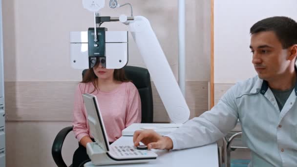 Οφθαλμολογική θεραπεία - νεαρή γυναίκα ελέγχει την οπτική οξύτητα σε οπτικό εξοπλισμό με γιατρό — Αρχείο Βίντεο