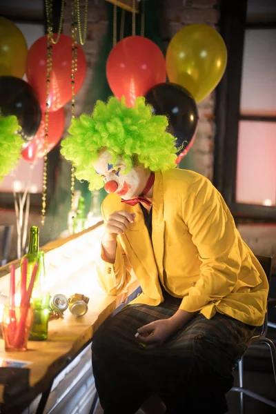 Clown concept - a tired sad man clown in yellow jacket and green wig sitting in the dressing room