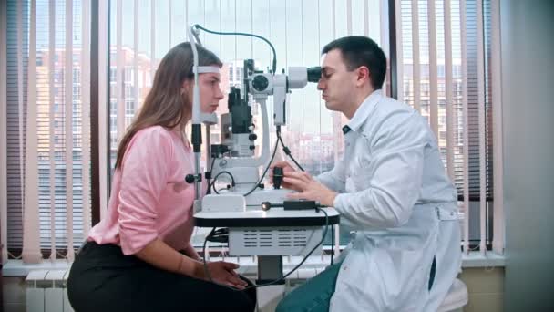 Tratamiento oftalmológico: un médico que revisa la agudeza visual de una mujer joven y bonita con un equipo especial en el espacioso gabinete en el fondo de la ventana panorámica — Vídeo de stock