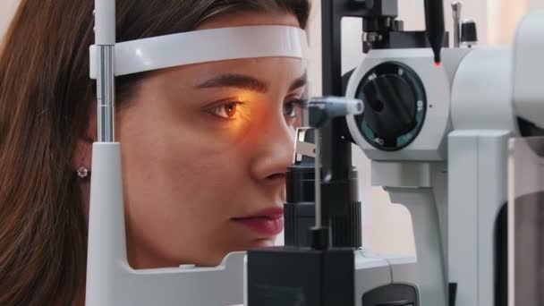 Ophthalmology treatment - a young woman with pink lips checking her visual acuity with a light of special big optometry machine - brown eyes color — Stock Video