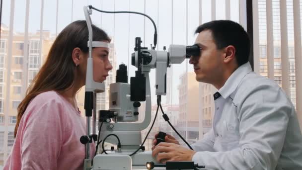 Oogheelkundige behandeling - een arts die jonge vrouwen controleert op gezichtsscherpte met een speciale machine - een ruime kast met panoramisch raam met rolluiken — Stockvideo