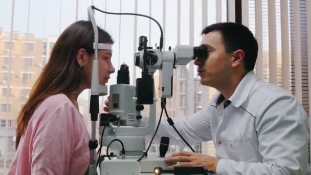 Ophthalmology treatment - a doctor checking young womans visual acuity with a big optometry machine - a spacious cabinet with panoramic window with blinds — Stockvideo