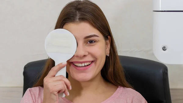 Tratamiento oftalmológico - joven mujer sonriente comprobando su agudeza visual - cerrando el ojo con un protector ocular y diciendo lo que dice la proyección — Foto de Stock