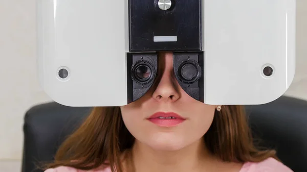 Tratamento oftalmológico - uma jovem mulher com lábios rosa brilhantes verificando sua acuidade visual com um equipamento especial de optometria — Fotografia de Stock