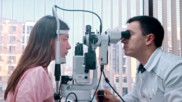 Οφθαλμολογική θεραπεία σε εσωτερικούς χώρους - ένας γιατρός τον έλεγχο των νέων γυναικών οπτική οξύτητα με ένα ειδικό μηχάνημα - ένα ευρύχωρο ντουλάπι με πανοραμική παράθυρο με περσίδες — Αρχείο Βίντεο