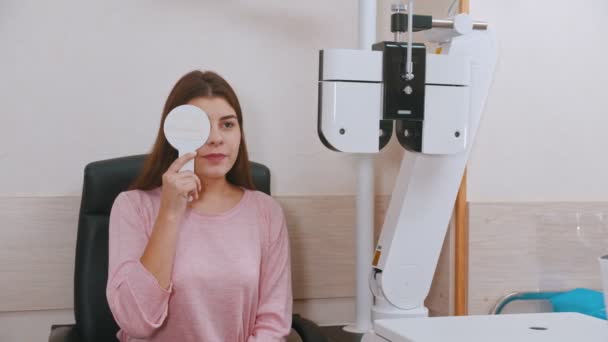 Oogheelkundige behandeling in het kabinet - jonge vrouw controleert haar gezichtsscherpte - sluit haar oog met een oogschild — Stockvideo