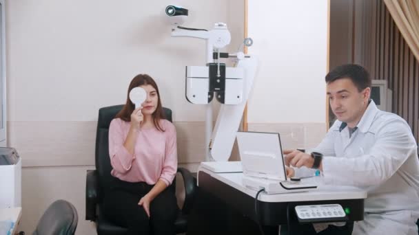 Oogheelkundige behandeling in het heldere kabinet - jonge vrouw controleert haar gezichtsscherpte - sluit haar oog met een oogschild en leest brieven — Stockvideo