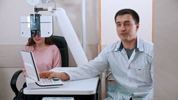 Офтальмологическое лечение в кабинете - молодая улыбающаяся женщина проверяет остроту зрения на оптическом оборудовании у врача — стоковое видео