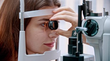Göz hekimliği tedavisi - pembe dudaklı genç bir kadın görme keskinliğini özel bir büyük optometri makinesi ve lens ışığı ile kontrol ediyor - kahverengi göz rengi