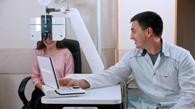 Oftalmoloji tedavisi - genç gülümseyen kadın görme keskinliğini optometrist ekipmanla kontrol ediyor - yanında oturan bir doktor