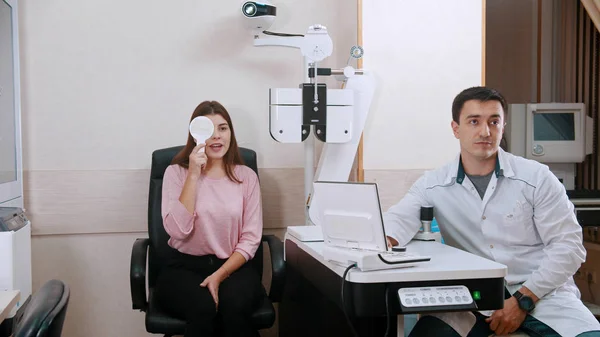 Augenheilkunde - junge Frau überprüft ihre Sehschärfe - Augen zu und Buchstaben lesen - Arzt sitzt in der Nähe — Stockfoto