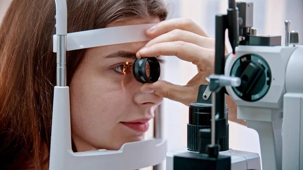 Tratamento oftalmológico - uma jovem mulher com lábios cor de rosa verificando sua acuidade visual com uma luz de grande máquina de optometria especial e lente - cor dos olhos castanhos — Fotografia de Stock