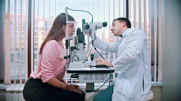 Oftalmologia - um médico verificando jovem mulher bonita acuidade visual com um equipamento especial no armário espaçoso no fundo da janela panorâmica — Fotografia de Stock