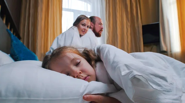 A family in the hotel room - a little girl sleeping in bed - her mom and dad looking at her — Stock Photo, Image