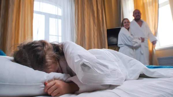 Rodina v hotelovém pokoji - holčička spící v posteli - její máma s tátou se na ni dívají a rozhrnou závěsy — Stock fotografie
