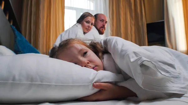 A family in the hotel room - a girl sleeping in bed - her mom and dad looking at her — Stock Photo, Image